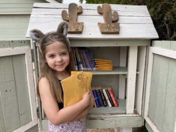 a young girl stands in front of a little library with her hands full of free art packets to distribute to her community