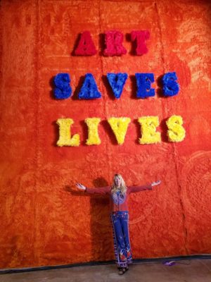 Lisa Rasmussen standing in front of an artwork that says Art Saves Lives.